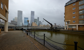 Canary Wharf Hotel Quay This urban photograph captures Canary Wharf Hotel Quay in London, United Kingdom, during the afternoon in early autumn. The image includes the waters of the quay with railings and a walkway in the foreground, while several modern high-rise buildings of Canary Wharf are clearly visible in the background across the Thames. Notable landmarks such as the One Canada Square tower and other recognisable skyscrapers add to the cityscape, alongside a historic quay crane. The photograph features the blend of contemporary architecture and dockside infrastructure typical of the Canary Wharf area in London.